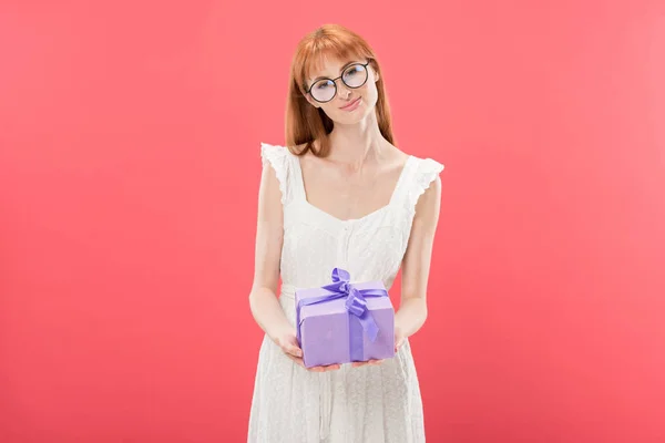 Vue de face de sourire rousse fille en lunettes et robe blanche tenant cadeau d'anniversaire et regardant la caméra isolée sur rose — Photo de stock