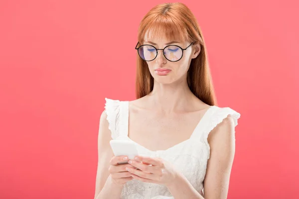 Mujer joven pelirroja disgustado en gafas y vestido blanco usando teléfono inteligente aislado en rosa - foto de stock