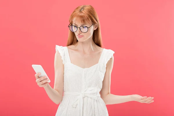 Mujer joven pelirroja disgustado en gafas y vestido blanco usando teléfono inteligente aislado en rosa - foto de stock