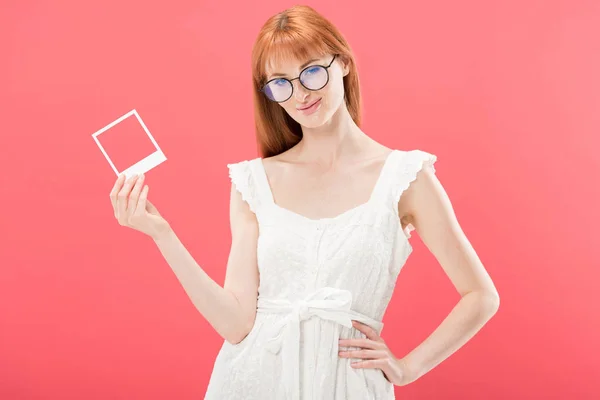 Pretty redhead young woman in glasses and white dress holding vintage camera frame and looking at camera isolated on pink — Stock Photo