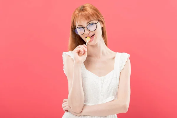 Attractive redhead young woman in glasses and white dress licking lollipop isolated on pink — Stock Photo