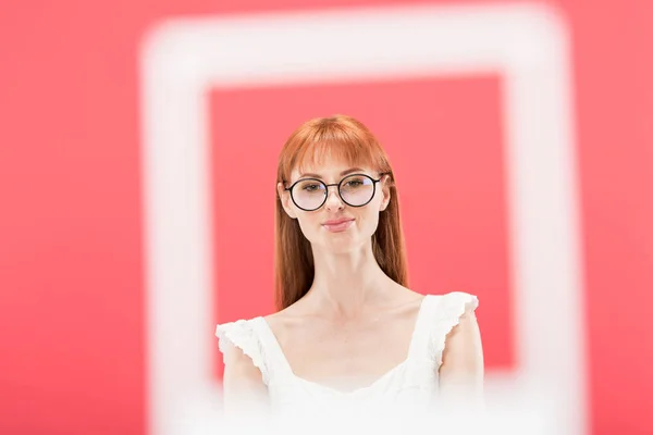 KYIV, UKRAINE - MAY 23, 2019: selective focus of redhead girl in glasses standing behind vintage camera frame isolated on pink — Stock Photo