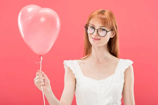 Усміхнена руда дівчина в окулярах і біла сукня тримає повітряну кулю у формі серця ізольовано на рожевому — стокове фото