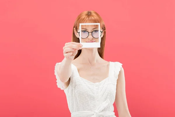 Front view of pretty redhead young woman in glasses and white dress holding vintage camera frame and looking at camera isolated on pink — Stock Photo
