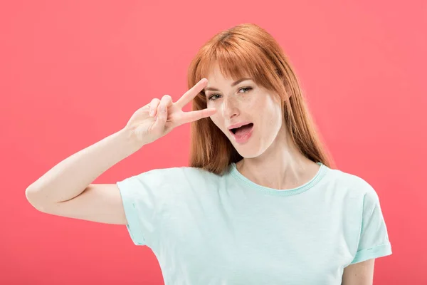 Attractive redhead girl in t-shirt smiling and showing peace sign isolated on pink — Stock Photo