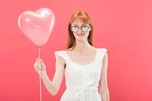 Front view of smiling redhead girl in glasses and white dress holding heart-shaped balloon isolated on pink — Stock Photo