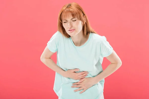 Mulher ruiva com dor de estômago tocando barriga com olhos fechados isolados em rosa — Fotografia de Stock