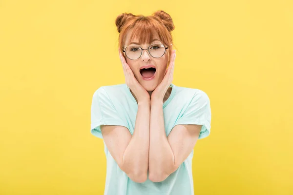 Frontansicht eines aufgeregten rothaarigen Mädchens mit Brille, das Gesicht berührt und isoliert auf gelb in die Kamera blickt — Stockfoto