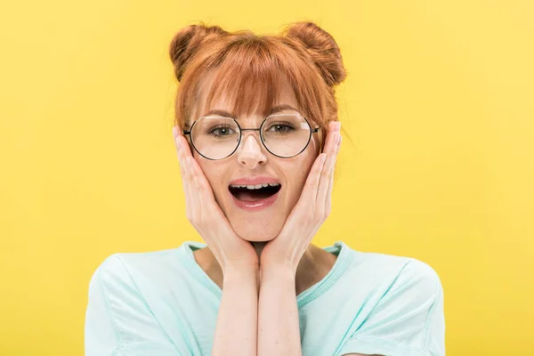 Front view of excited redhead girl in glasses touching face and looking at camera isolated on yellow — Stock Photo