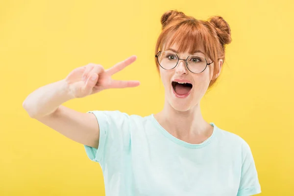 Excited redhead girl in glasses and t-shirt showing peace sign isolated on yellow — Stock Photo