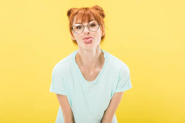 Vista frontale della ragazza rossa allegra in occhiali sporgenti lingua e guardando la fotocamera isolata sul giallo — Foto stock