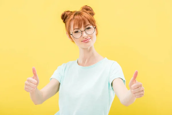 Visão frontal da menina ruiva sorridente em óculos mostrando polegares para cima e olhando para a câmera isolada no amarelo — Fotografia de Stock