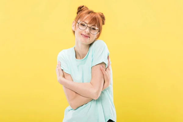 Pleased redhead girl in glasses and t-shirt embracing herself and looking at camera with smile isolated on yellow — Stock Photo
