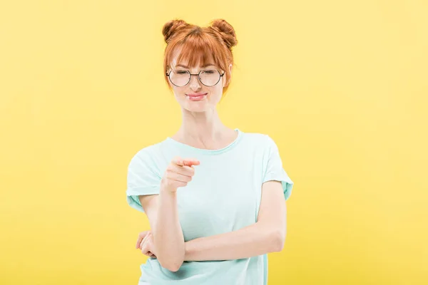 Vista frontale di ragazza rossa sorridente in occhiali e t-shirt che punta con il dito alla fotocamera isolata sul giallo — Foto stock