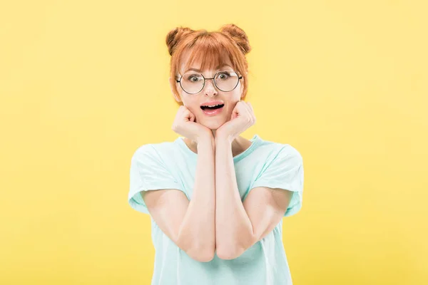 Vista frontal de chica pelirroja sorprendida en gafas apuntalando la cara con las manos y mirando la cámara aislada en amarillo - foto de stock