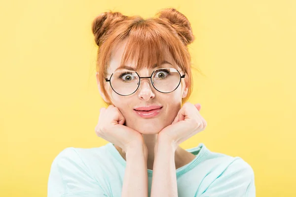 Front view of surprised redhead girl in glasses propping face with hands and looking at camera isolated on yellow — Stock Photo