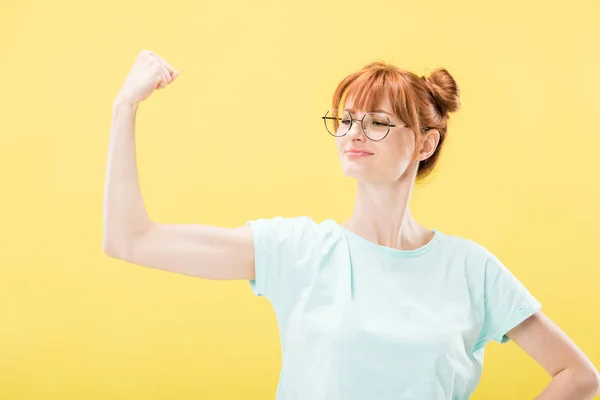 Confident pleased redhead girl in glasses and t-shirt holding fist up isolated on yellow — Stock Photo