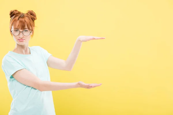 Attraktives rothaariges Mädchen in T-Shirt und Brille, das mit den Händen zeigt und isoliert auf gelb in die Kamera schaut — Stockfoto