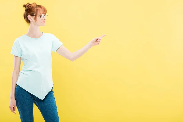 Rossa ragazza in occhiali e t-shirt punta con dito isolato su giallo — Foto stock