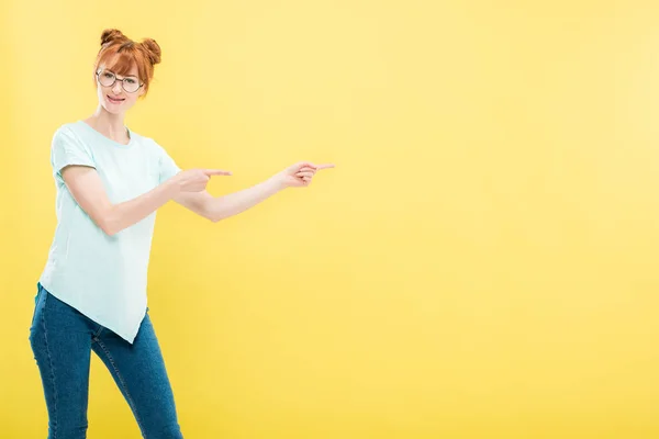 Smiling redhead girl in glasses and t-shirt pointing with fingers and looking at camera isolated on yellow — Stock Photo
