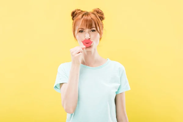 Front view of redhead girl in t-shirt holding paper lips and looking at camera isolated on yellow — Stock Photo