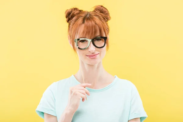Vue de face de fille rousse souriante en t-shirt tenant des lunettes de jouet et regardant la caméra isolée sur jaune — Photo de stock