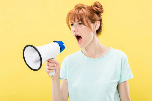 Сердитая рыжая молодая женщина, держащая мегафон и кричащая изолированно на желтом — стоковое фото