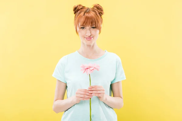 Vista frontale di rossa giovane donna in t-shirt con fiore in mano e guardando la fotocamera con sorriso isolato sul giallo — Foto stock