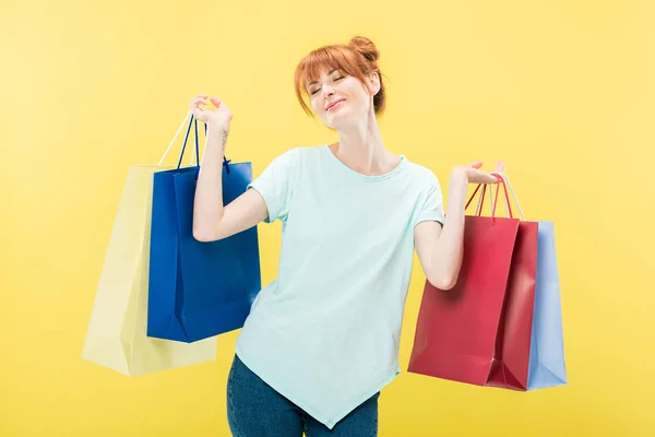 Sorridente rossa ragazza che tiene borse della spesa con gli occhi chiusi isolati sul giallo — Foto stock