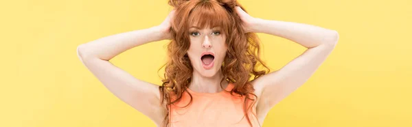 Plan panoramique de rousse bouclée choquée femme touchant les cheveux et regardant la caméra isolée sur jaune — Photo de stock