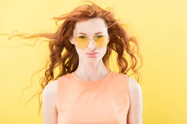 Вид спереди серьезной кудрявой рыжеволосой женщины в солнцезащитных очках, изолированных на желтом — стоковое фото