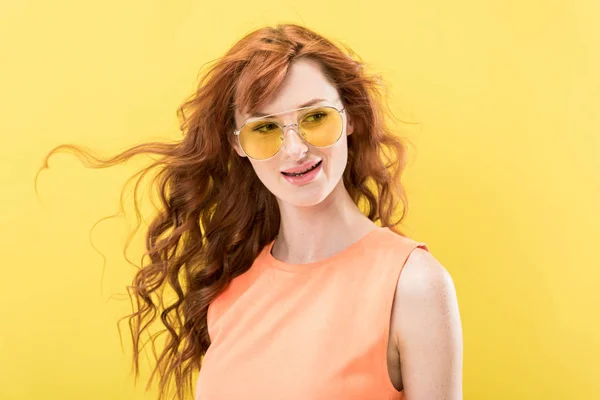 Довольно кудрявая рыжая женщина в солнечных очках смотрит в сторону изолированных на желтый — стоковое фото