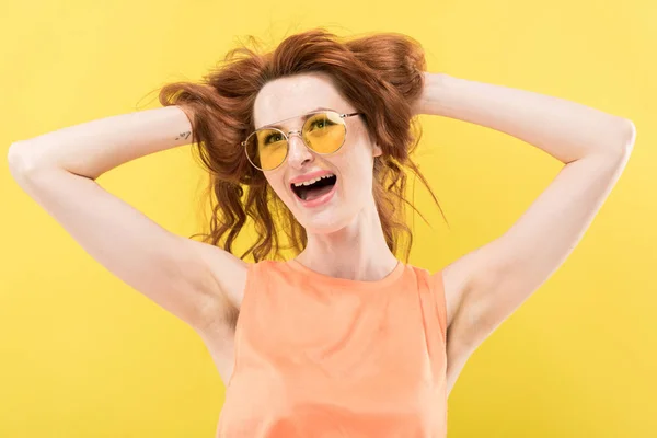 Збуджена руда дівчина в сонцезахисних окулярах торкається кучерявого волосся ізольовано на жовтому — стокове фото