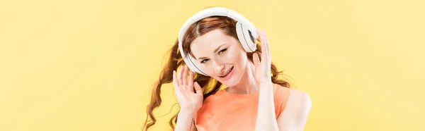 Panoramic shot of attractive redhead woman listening music in headphones and smiling isolated on yellow — Stock Photo