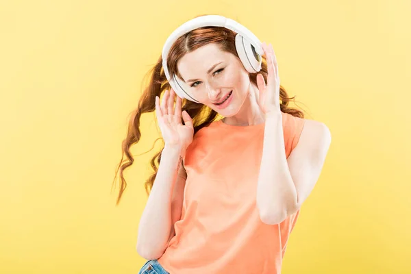 Atractiva pelirroja escuchando música en auriculares y sonriendo aislada en amarillo - foto de stock