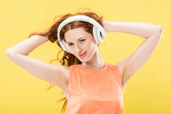 Attractive redhead woman listening music in headphones and touching curly hair isolated on yellow — Stock Photo