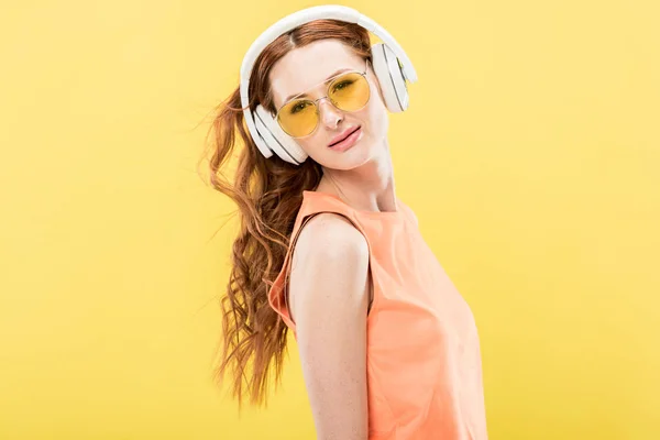 Atractiva pelirroja en gafas de sol escuchando música en auriculares aislados en amarillo - foto de stock