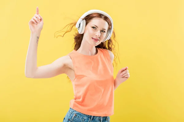 Atractiva pelirroja sonriente escuchando música en auriculares y bailando aislada en amarillo - foto de stock