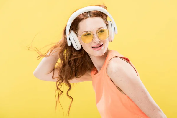 Atractiva pelirroja en gafas de sol escuchando música en auriculares y sonriendo aislada en amarillo - foto de stock