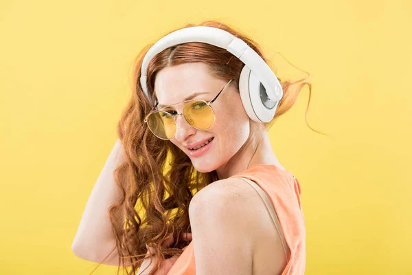 Atractiva pelirroja en gafas de sol escuchando música en auriculares y sonriendo aislada en amarillo - foto de stock