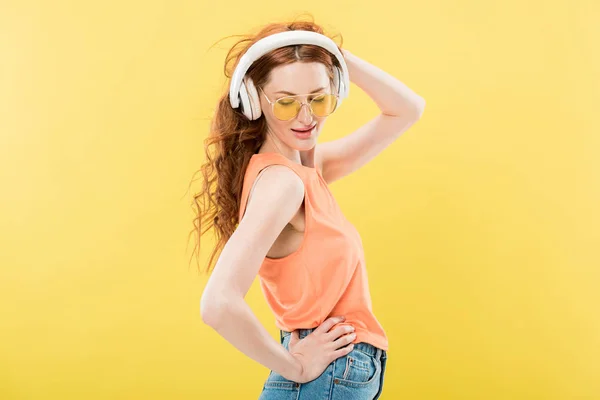 Приваблива руда жінка в сонцезахисних окулярах слухає музику в навушниках і посміхається ізольовано на жовтому — стокове фото