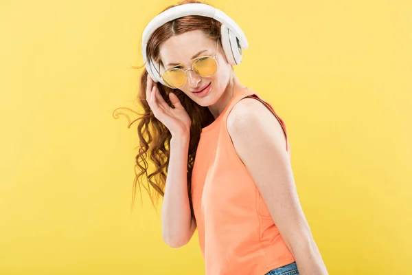 Attraente rossa donna in occhiali da sole ascoltare musica in cuffia e sorridente isolato su giallo — Foto stock