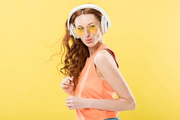 Jolie rousse femme en lunettes de soleil écouter de la musique dans les écouteurs isolés sur jaune — Photo de stock