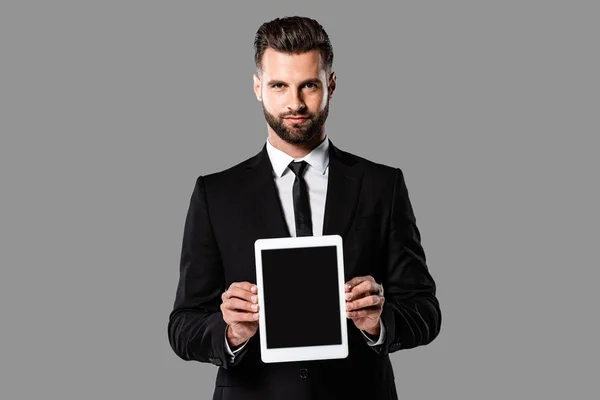 Guapo hombre de negocios en traje negro mostrando tableta digital con pantalla en blanco aislado en gris - foto de stock