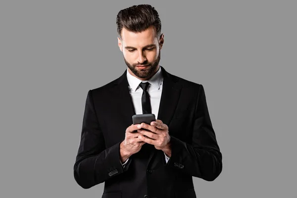 Hombre de negocios barbudo en traje negro usando teléfono inteligente aislado en gris - foto de stock