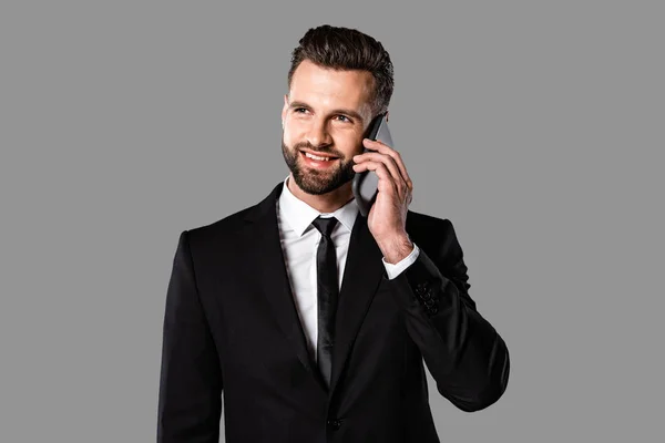 Handsome smiling businessman in black suit talking on smartphone isolated on grey — Stock Photo