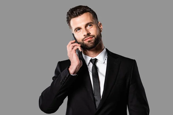 Soñador guapo hombre de negocios en traje negro hablando en el teléfono inteligente aislado en gris - foto de stock