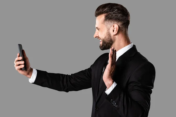 Vista lateral de empresário sorridente em terno preto ter chamada de vídeo no smartphone e mão acenando isolado em cinza — Fotografia de Stock