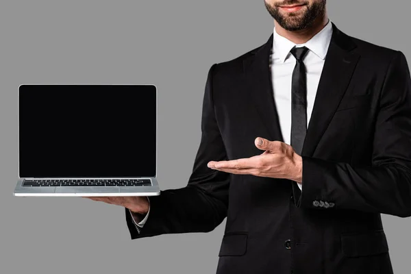 Vista cortada de empresário em terno preto apontando com a mão para laptop com tela em branco isolado em cinza — Fotografia de Stock