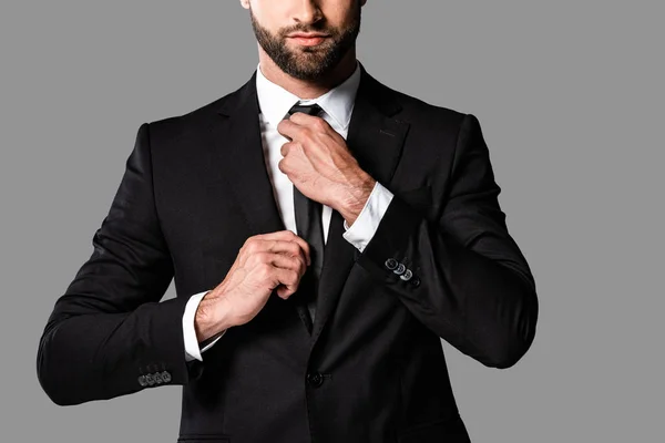 Vista recortada de elegante hombre de negocios guapo en traje negro fijación corbata aislado en gris - foto de stock